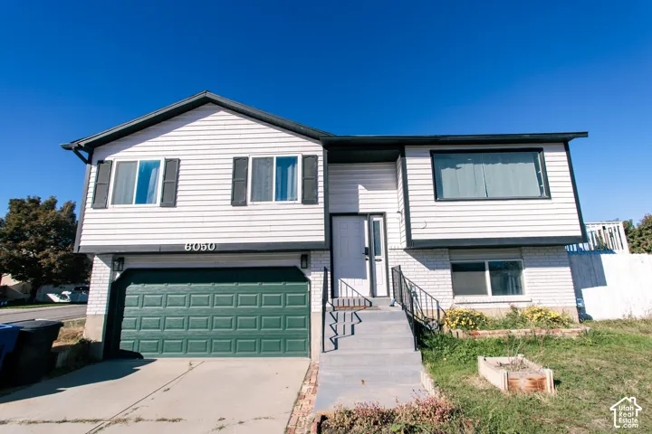 Split-level exterior featuring light-toned horizontal siding, a lower-level masonry facade, an attached garage with a sectional door, and metal handrails along the concrete entry stairs
