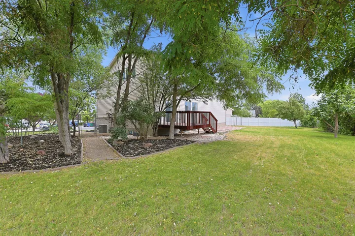 Expansive backyard featuring a manicured lawn, established trees, a wood-finish deck with stairs, a gravel path, and a white privacy fence