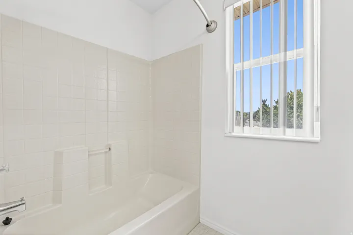 Bathroom featuring a white bathtub with integrated shelving, tiled shower surround, and a window with vertical blinds