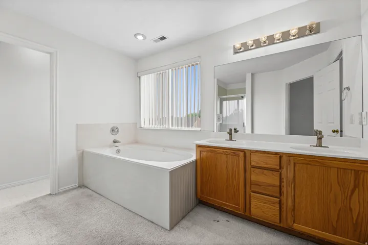 Bathroom featuring a dual vanity with wood cabinetry, a built-in soaking tub, a large wall-mounted mirror, and a window with horizontal blinds
