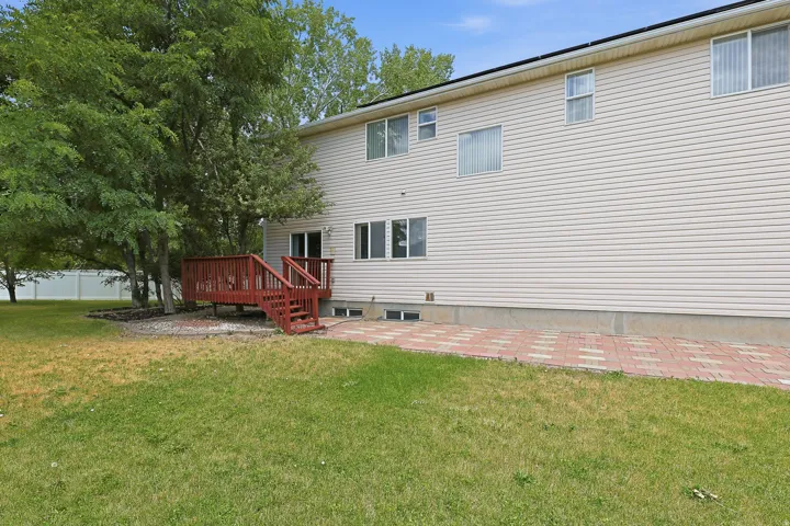 Rear exterior featuring a red-stained wooden deck with stairs to the yard, light-colored horizontal siding, multiple windows, and a brick paver patio