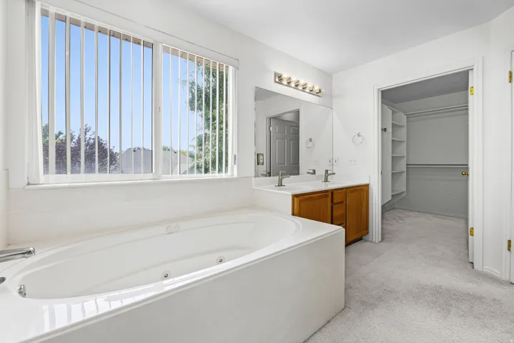 Spa-like bathroom featuring a jetted soaking tub, dual vanity with wood-finish cabinetry, and a spacious walk-in closet with built-in shelving