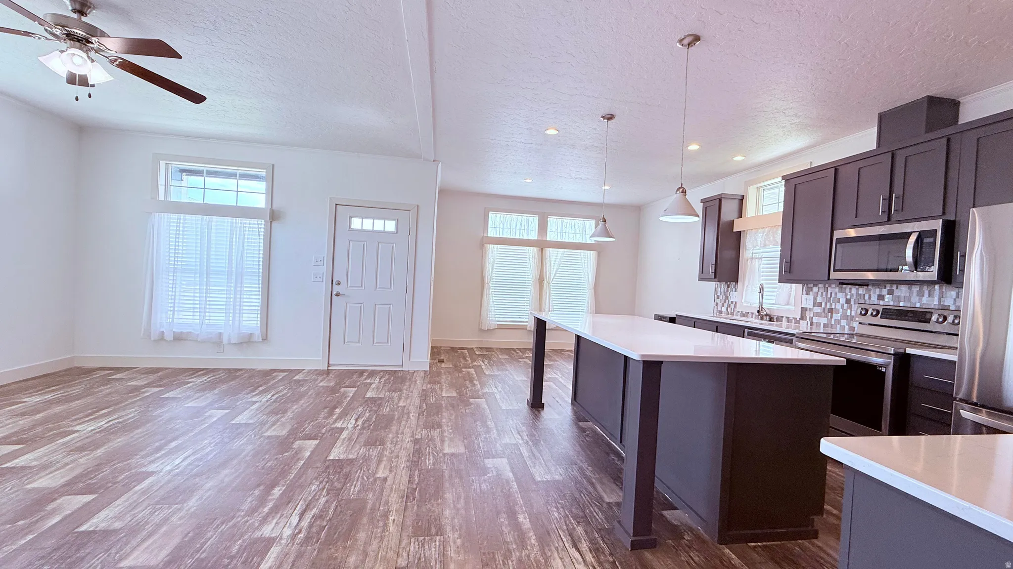 Open-concept living space featuring laminate flooring, a contemporary kitchen island with pendant lighting, gray cabinetry, stainless steel appliances, and backsplash