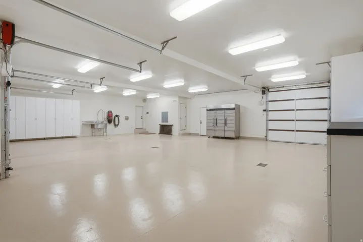 Spacious garage featuring a light-toned epoxy floor finish, abundant overhead fluorescent lighting, extensive built-in white cabinetry, a stainless steel utility sink, and two panel garage doors
