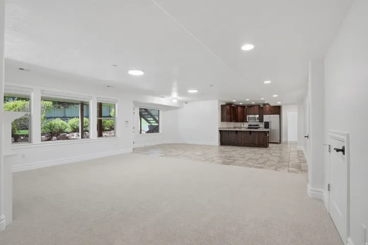 Spacious lower-level living area featuring large windows, light-toned carpeting, recessed lighting, and a built-in kitchenette with dark wood cabinetry, a stainless steel refrigerator, and a tiled floor