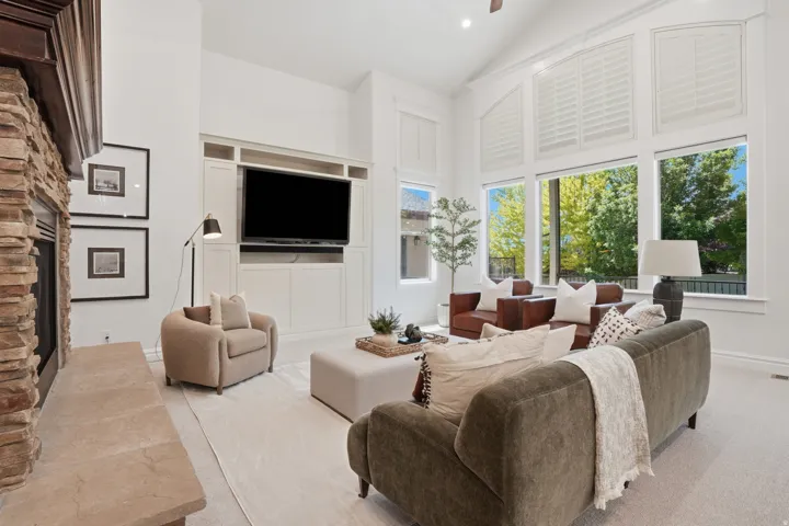 Spacious living area featuring a stone-clad fireplace with a natural stone hearth, built-in media cabinetry, soaring vaulted ceilings, and extensive window-frontage with upper plantation shutters