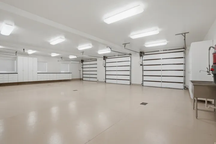 Spacious multi-car garage featuring a light-toned epoxy floor, multiple overhead fluorescent light fixtures, three sectional garage doors, extensive built-in white cabinetry, and a utility sink