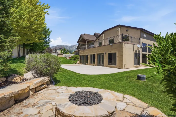 Rear exterior featuring a large concrete patio, multiple sliding glass doors, upper-level balcony with metal railing, and a flagstone fire pit area with stone seating