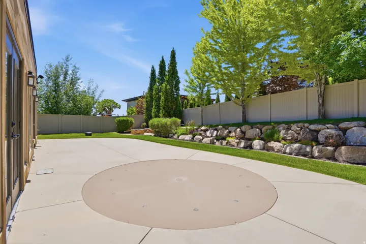 Spacious patio featuring a concrete surface with a circular inset, bordered by green lawn and a rock retaining wall
