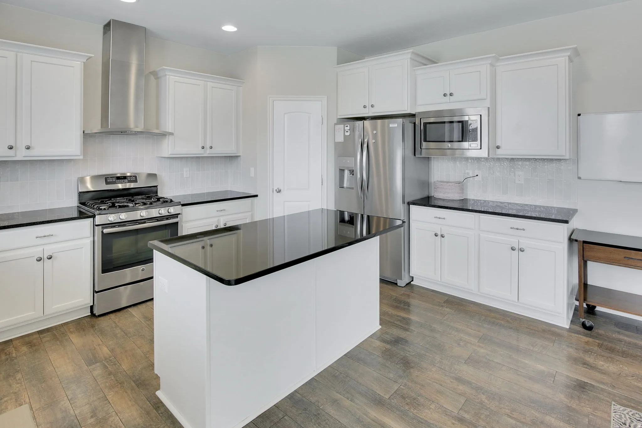 Kitchen featuring a central island with a black countertop, stainless steel appliances, white cabinetry with silver hardware, a tile backsplash, and wood-finish flooring