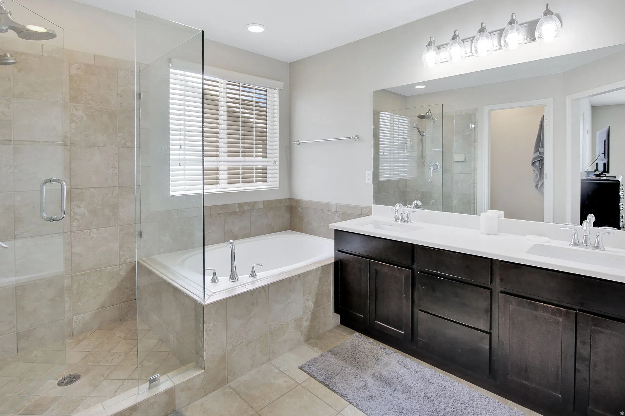 Spacious bathroom featuring a glass-enclosed shower, built-in soaking tub, dual vanities with white countertops, dark wood-finish cabinetry, and tiled flooring