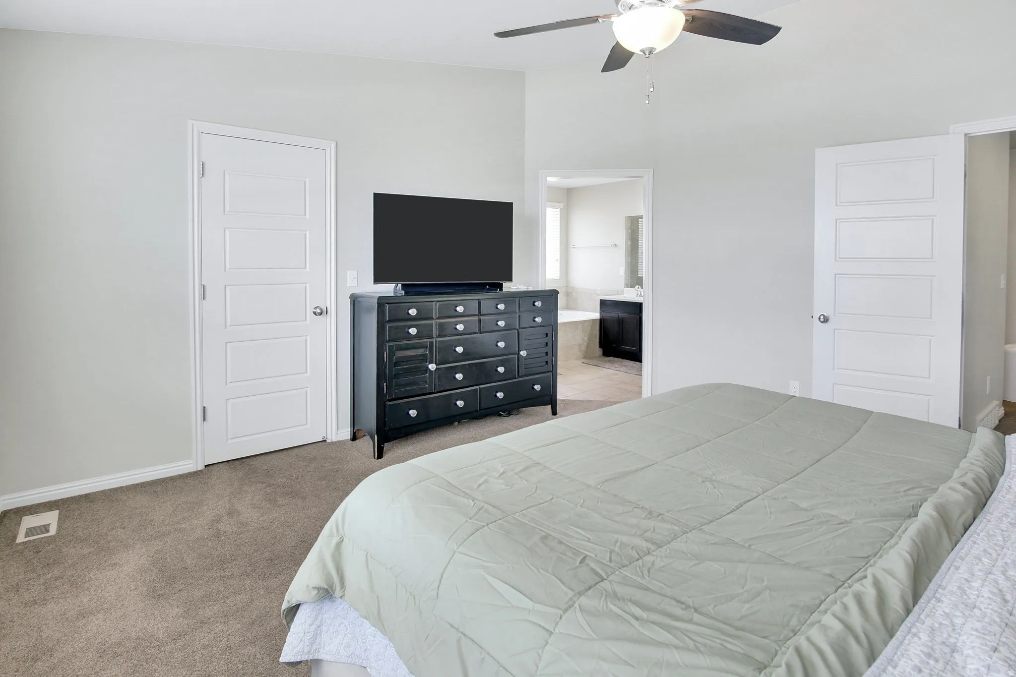 Carpeted bedroom featuring a ceiling fan with integrated lighting, two five-panel interior doors, and a partial view of an adjacent bathroom with a built-in tub and vanity