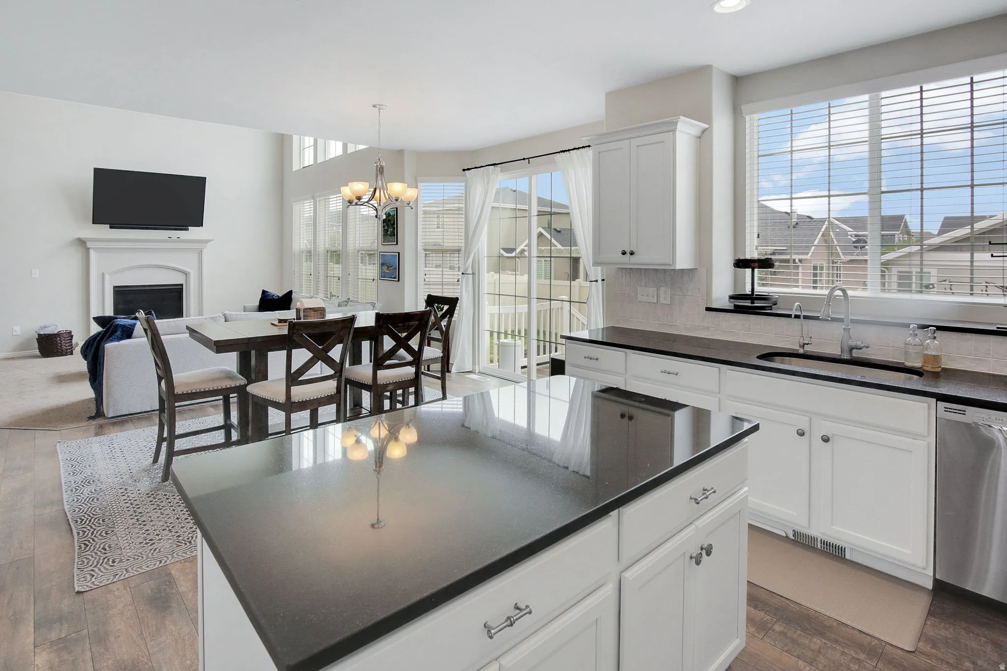 Open-concept living space featuring wood-finish flooring, a kitchen island with dark countertops, white cabinetry, a large window over the sink, and a fireplace with a white mantel