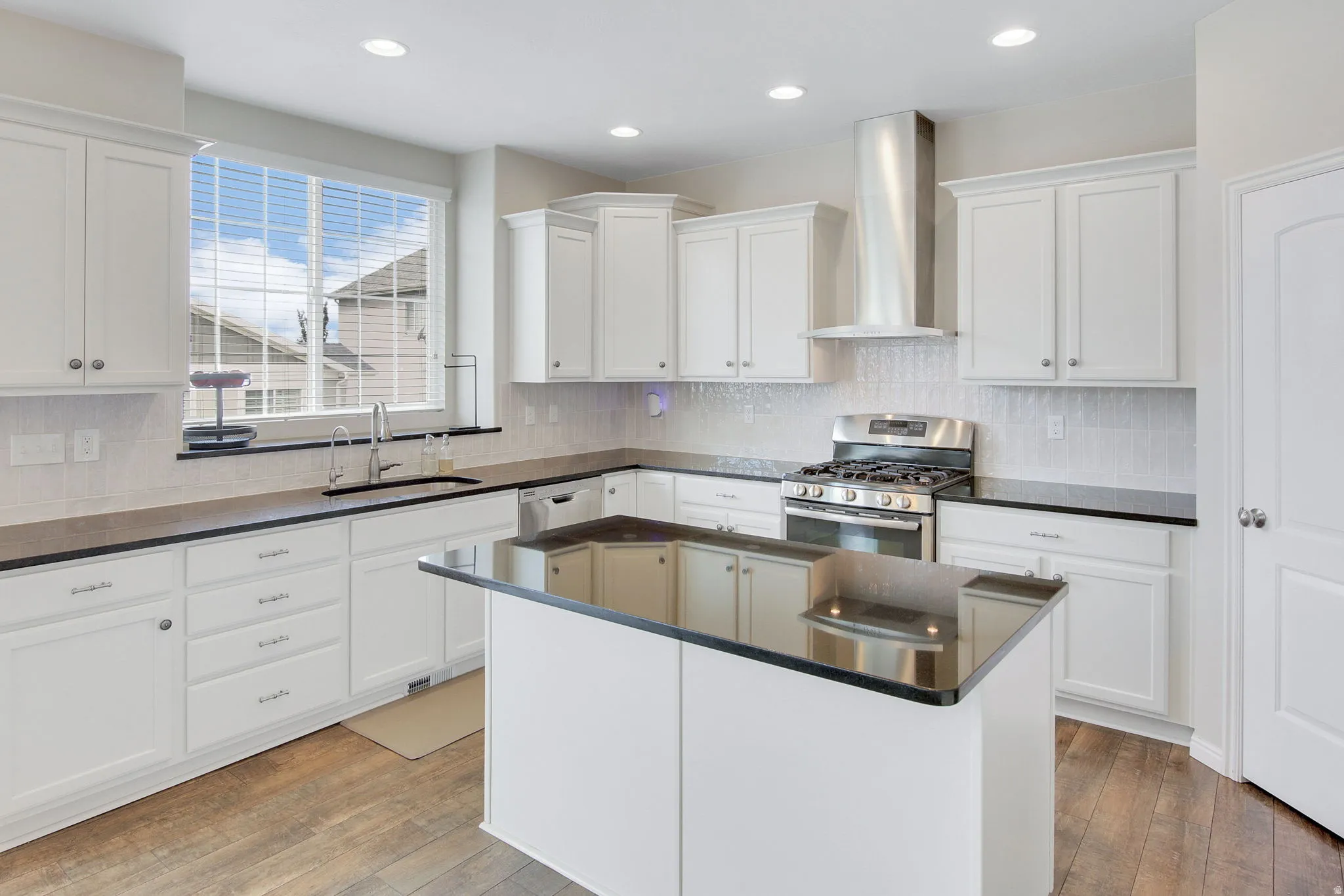 Contemporary kitchen featuring an island with a black countertop, white shaker-style cabinetry, stainless steel range with matching vent hood, recessed lighting, and wood-finish flooring