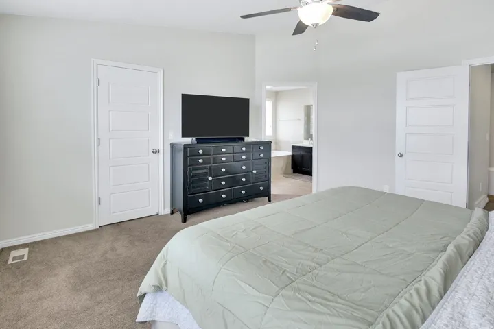Carpeted bedroom featuring a ceiling fan with integrated lighting, two five-panel interior doors, and a partial view of an adjacent bathroom with a built-in tub and vanity