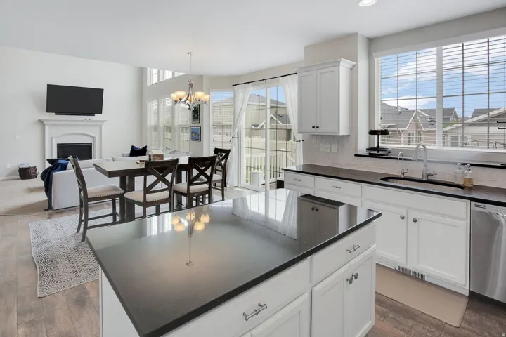 Open-concept living space featuring wood-finish flooring, a kitchen island with dark countertops, white cabinetry, a large window over the sink, and a fireplace with a white mantel