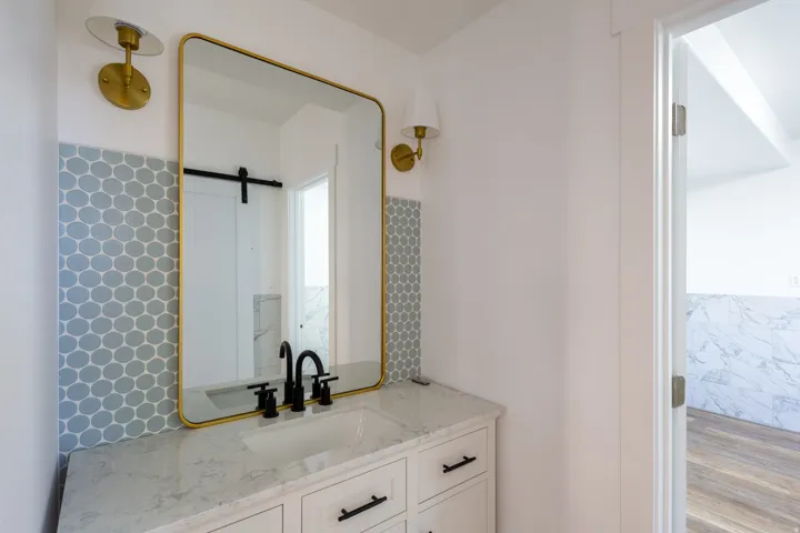 Vanity featuring a gold-framed mirror, dual brass sconces, hexagon tile backsplash, and a white countertop with an integrated sink