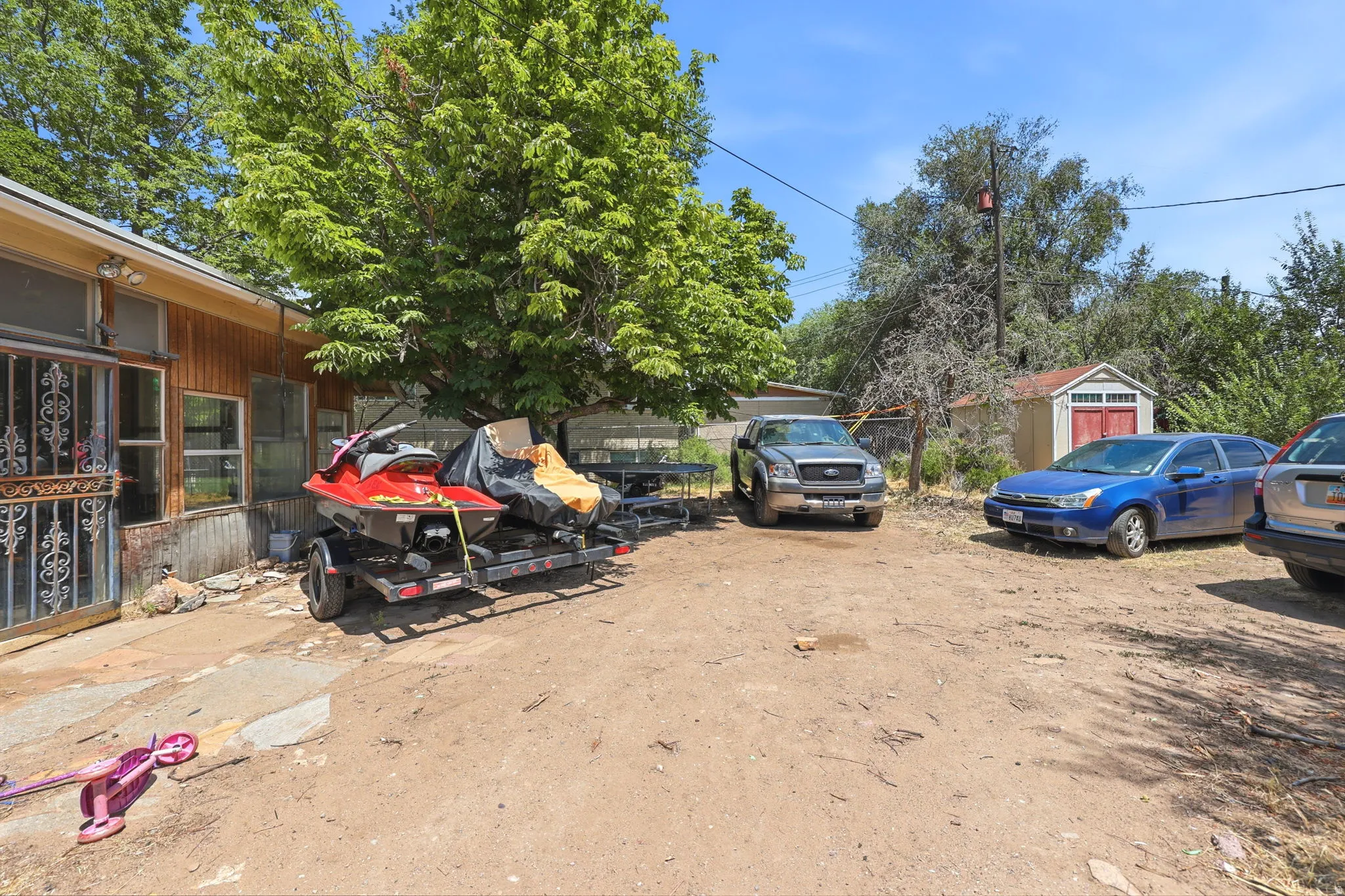 Expansive dirt lot providing ample outdoor parking, featuring a rustic wooden shed with red double doors, and mature trees providing natural shade