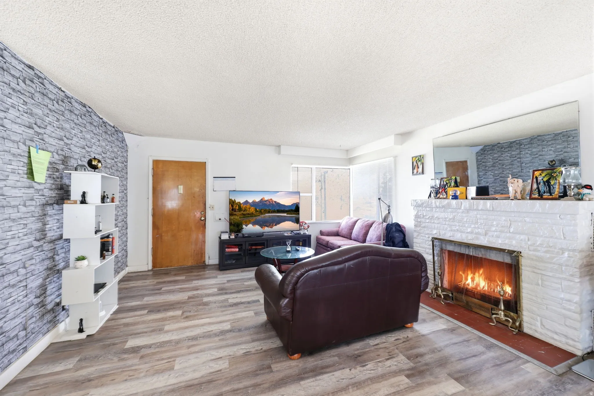 Living area with wood-finish flooring, a white-painted brick fireplace, an exposed stone-look accent wall, and a wood entry door