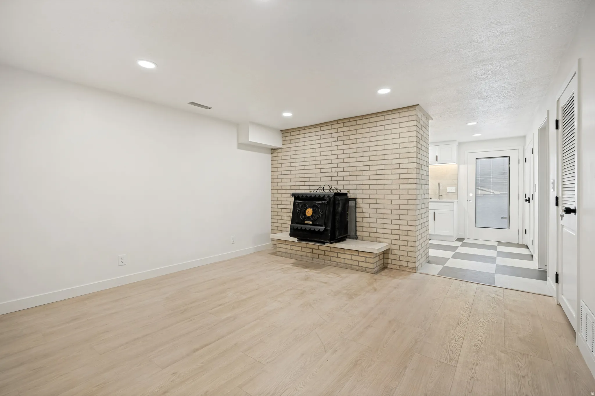 Spacious room featuring light wood-finish flooring, recessed lighting, and a prominent brick-clad fireplace with a black stove insert