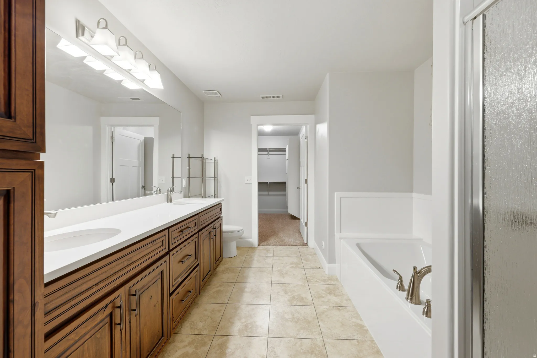 Bathroom featuring a dual vanity with wood cabinetry and white countertops, a built-in bathtub with brushed nickel fixtures, a glass-enclosed shower, and tile flooring