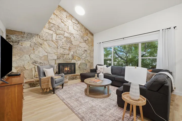 Vaulted ceiling living area featuring a full-height stone accent wall, an integrated fireplace, expansive window, a circular skylight, and light wood-finish flooring