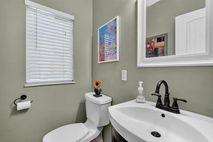 Powder room featuring a white single-hung window with blinds, a white rectangular mirror, and a white integrated sink with a dark bronze faucet