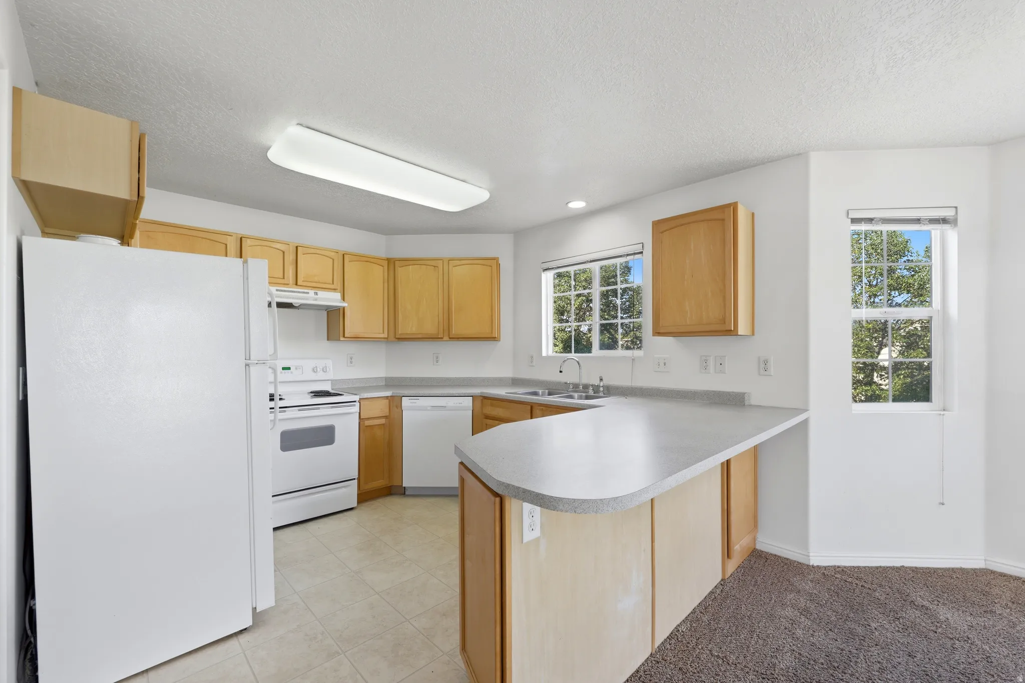 Kitchen featuring light wood cabinetry, white appliances, a peninsula with a light-toned countertop, tile flooring, and multiple windows