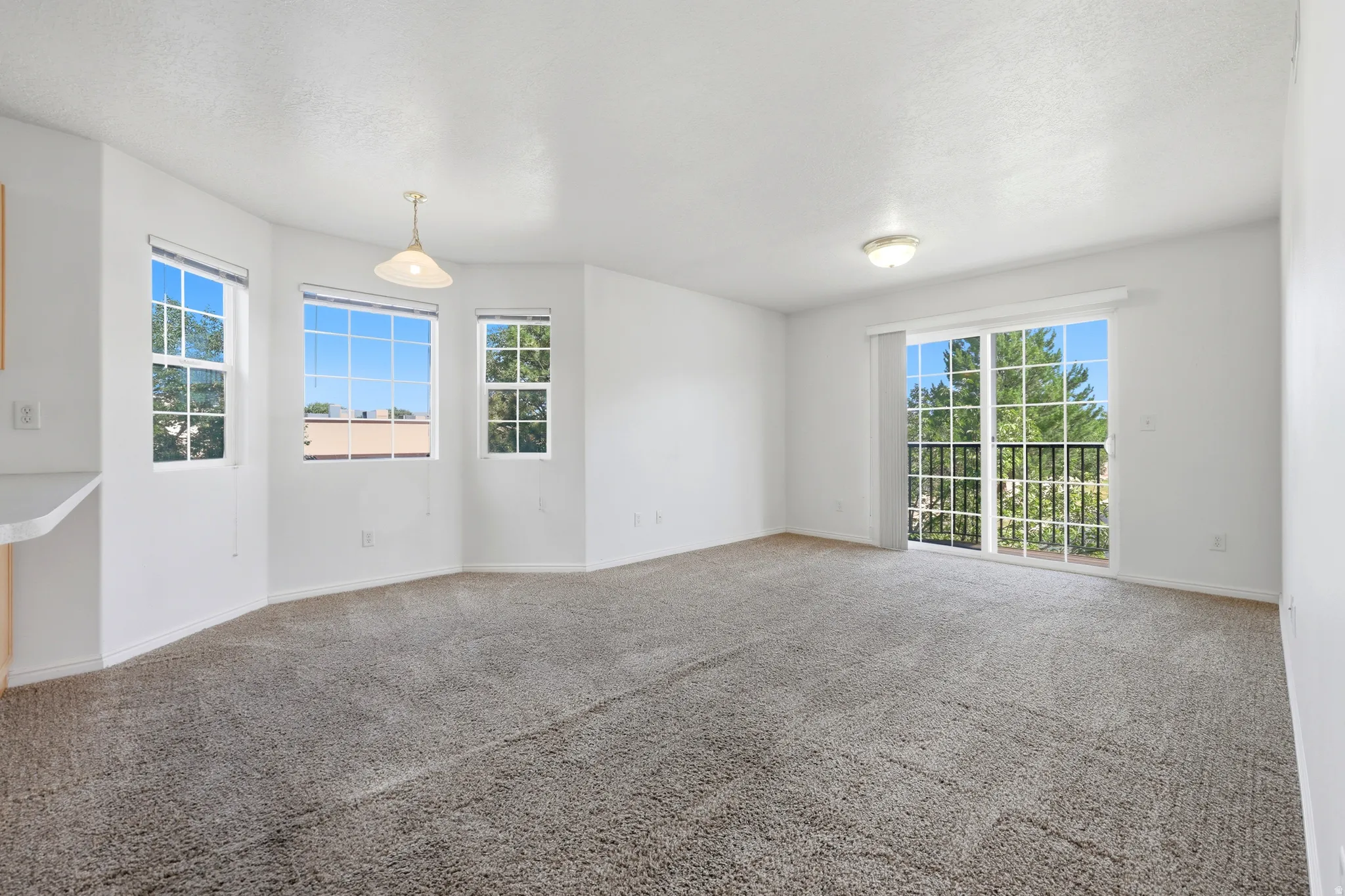 Spacious living area featuring light gray carpet, multiple windows, a sliding glass door with balcony access, a ceiling-mounted light fixture, and a pendant light