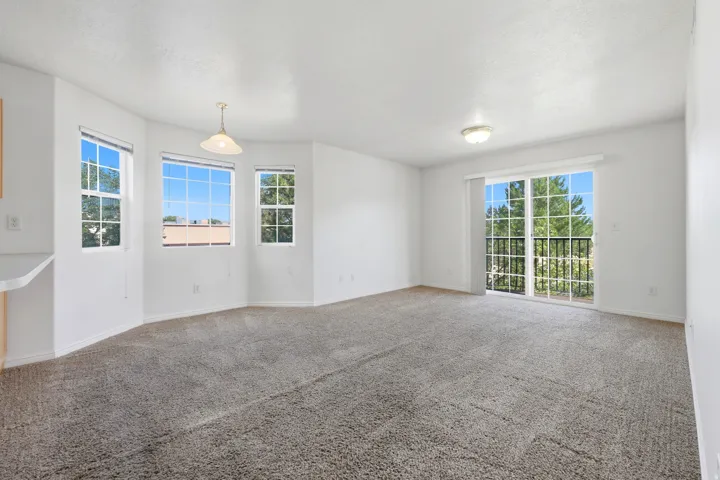Spacious living area featuring light gray carpet, multiple windows, a sliding glass door with balcony access, a ceiling-mounted light fixture, and a pendant light
