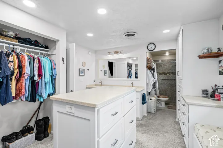 Spacious dressing area featuring a central island with a light-toned countertop, dual vanity sinks, extensive built-in cabinetry, and a walk-in shower with tiled walls