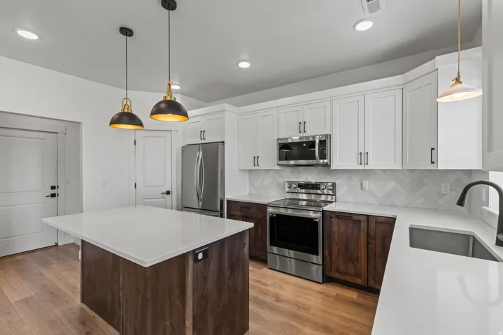 Contemporary kitchen featuring a large central island with a white countertop, two-tone cabinetry, stainless steel appliances, a herringbone tile backsplash, and wood-finish flooring