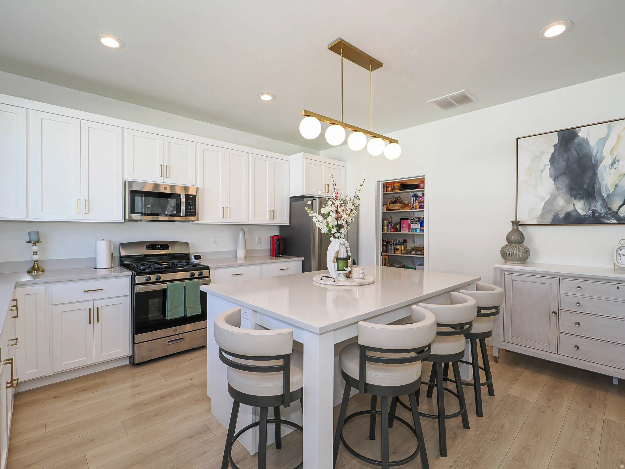 Modern kitchen featuring a central island with seating for four, white shaker-style cabinetry, stainless steel appliances, wood-finish flooring, and a walk-in pantry