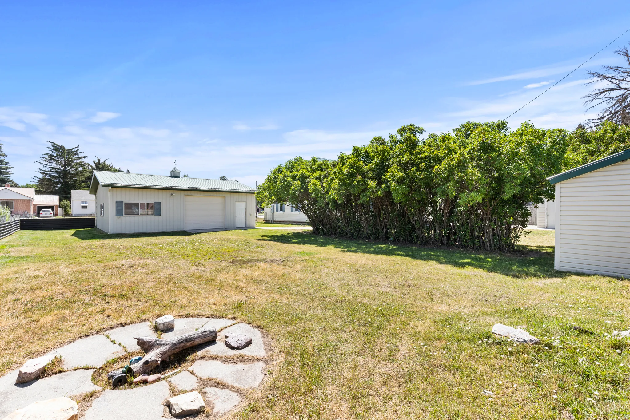 Expansive grassy yard featuring a large detached garage with a green metal roof, a second white siding outbuilding, and a row of mature green shrubs