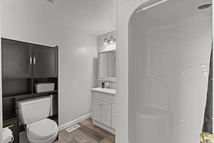 Compact bathroom featuring a white single-piece shower, dark wood-finish storage cabinetry with gold-tone hardware, a white vanity with under-sink storage, a mirrored medicine cabinet, and wood-finish flooring