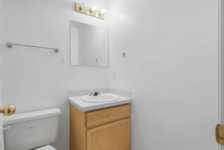 Clean and functional bathroom featuring a wood-finish vanity with integrated sink, white ceramic countertop, framed mirror with integrated lighting, white porcelain toilet, and brushed metal hardware and towel bar