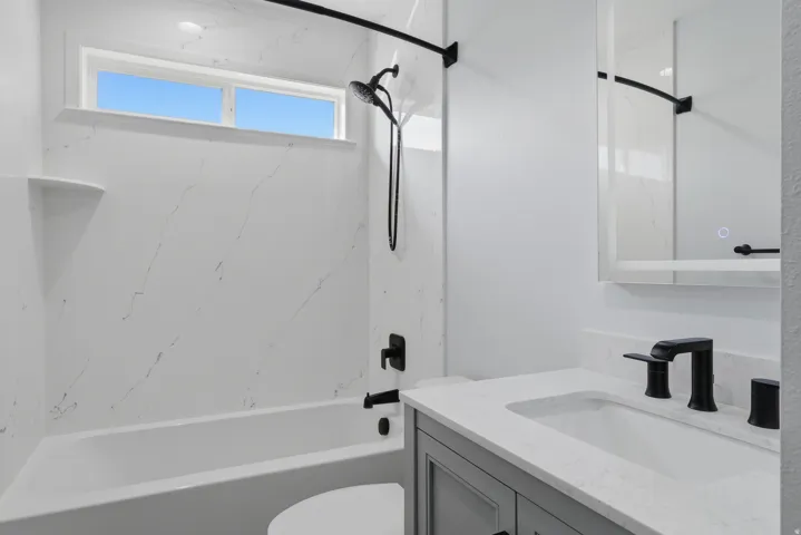 Bathroom featuring a white tub with a shower, matte black fixtures, a window above the tub, a vanity with a white countertop, and a wall-mounted mirror