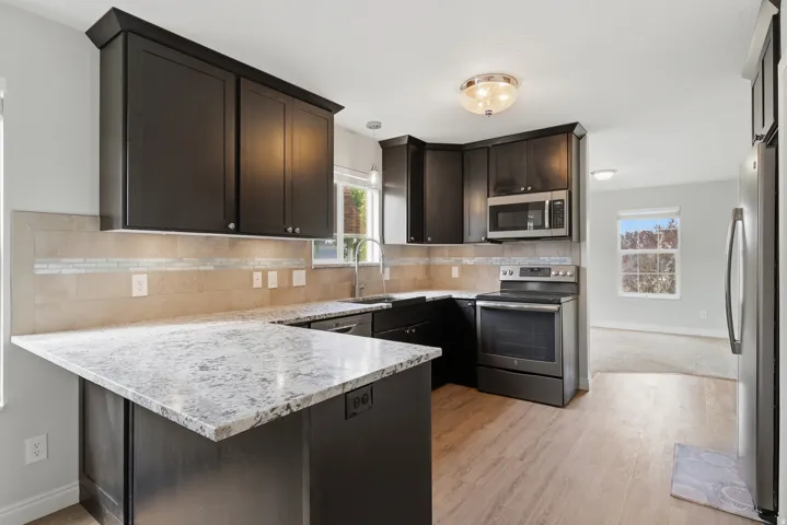 Modern kitchen featuring dark cabinetry with ample storage, a large marble-finish island with seating, stainless steel appliances including a built-in microwave and electric range, light tile backsplash, and wood-finish flooring throughout the open layout