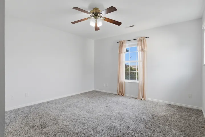 Spacious bedroom with neutral wall finishes, plush carpeting, ceiling fan with integrated lighting, and large windows framed with curtain rods for natural light and privacy control