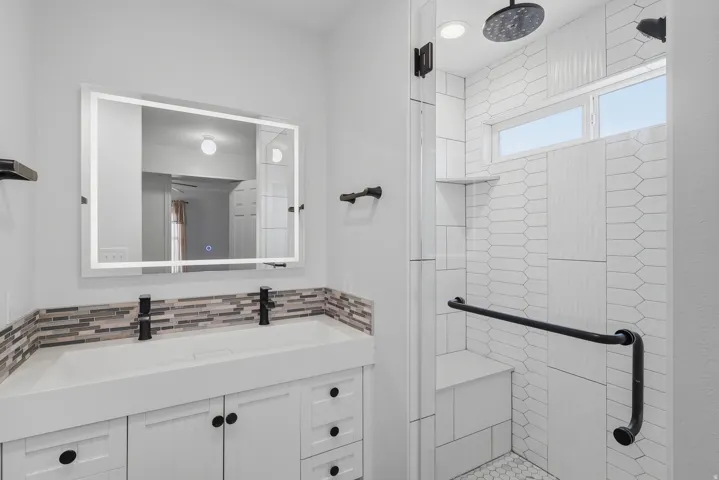 Dual sink vanity with integrated countertop and matte black faucets, illuminated mirror, walk-in shower with textured tile, built-in shower bench, and rainfall shower head