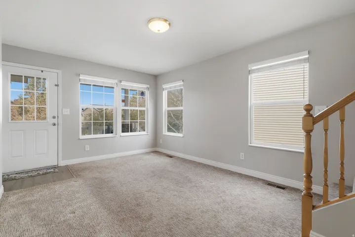 Bright foyer with multiple windows framing natural light, neutral gray walls, neutral carpet flooring, and a wooden stair railing