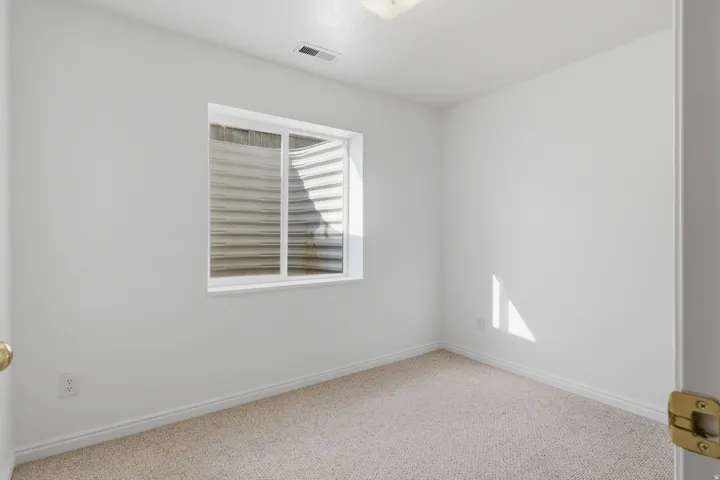 Bright, neutral bedroom featuring carpet flooring, a large window with plantation shutters providing natural light, and clean white walls with brass hardware accents