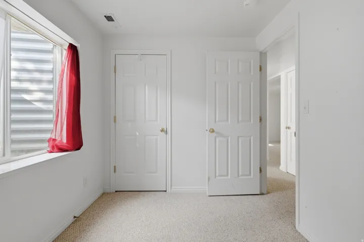 Bright hallway featuring multiple six-panel closet doors with brass hardware, neutral carpet flooring, and abundant natural light from a large window with red fabric treatment