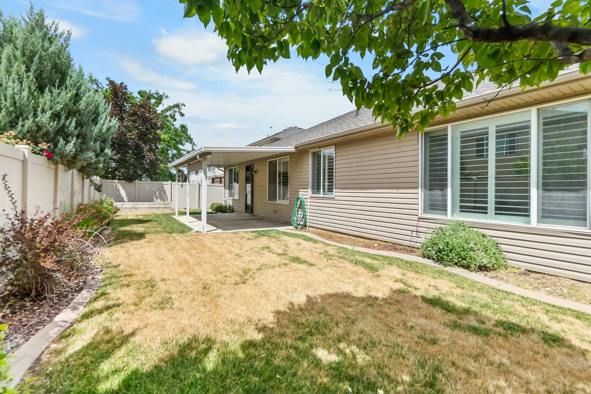 Expansive backyard featuring a covered patio, concrete hardscaping, vinyl fencing, mature landscaping, and bay windows with plantation shutters