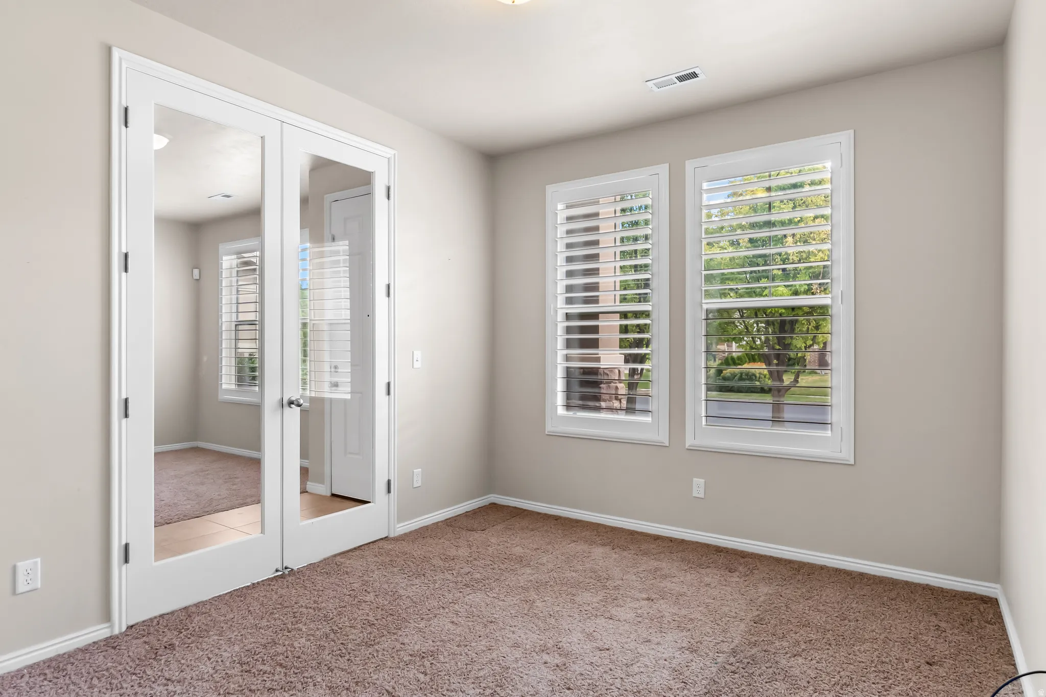 Room with white French doors, two windows featuring white louvered shutters, light beige wall paint, and brown carpet flooring