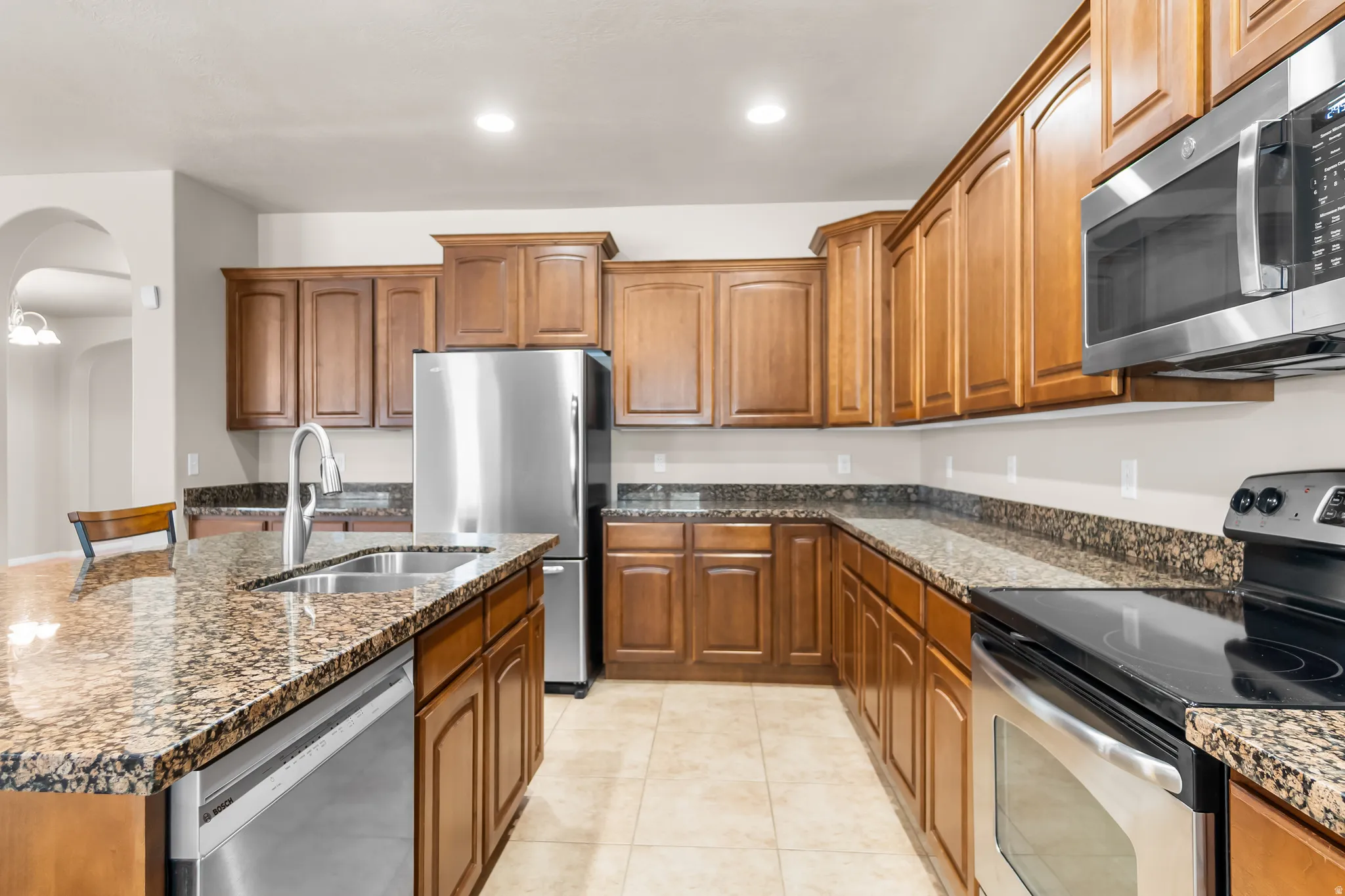 Spacious kitchen featuring warm wood cabinetry, speckled countertops, stainless steel appliances, and a central island with an integrated sink