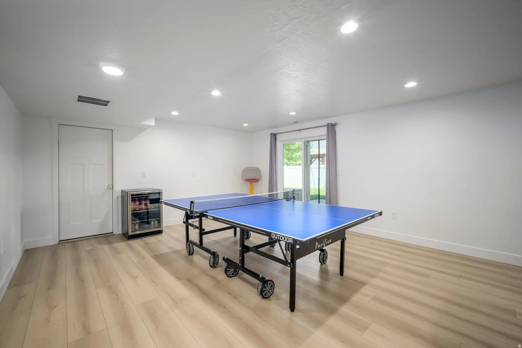 Spacious recreation room featuring light wood-finish flooring, recessed ceiling lights, and crisp white walls