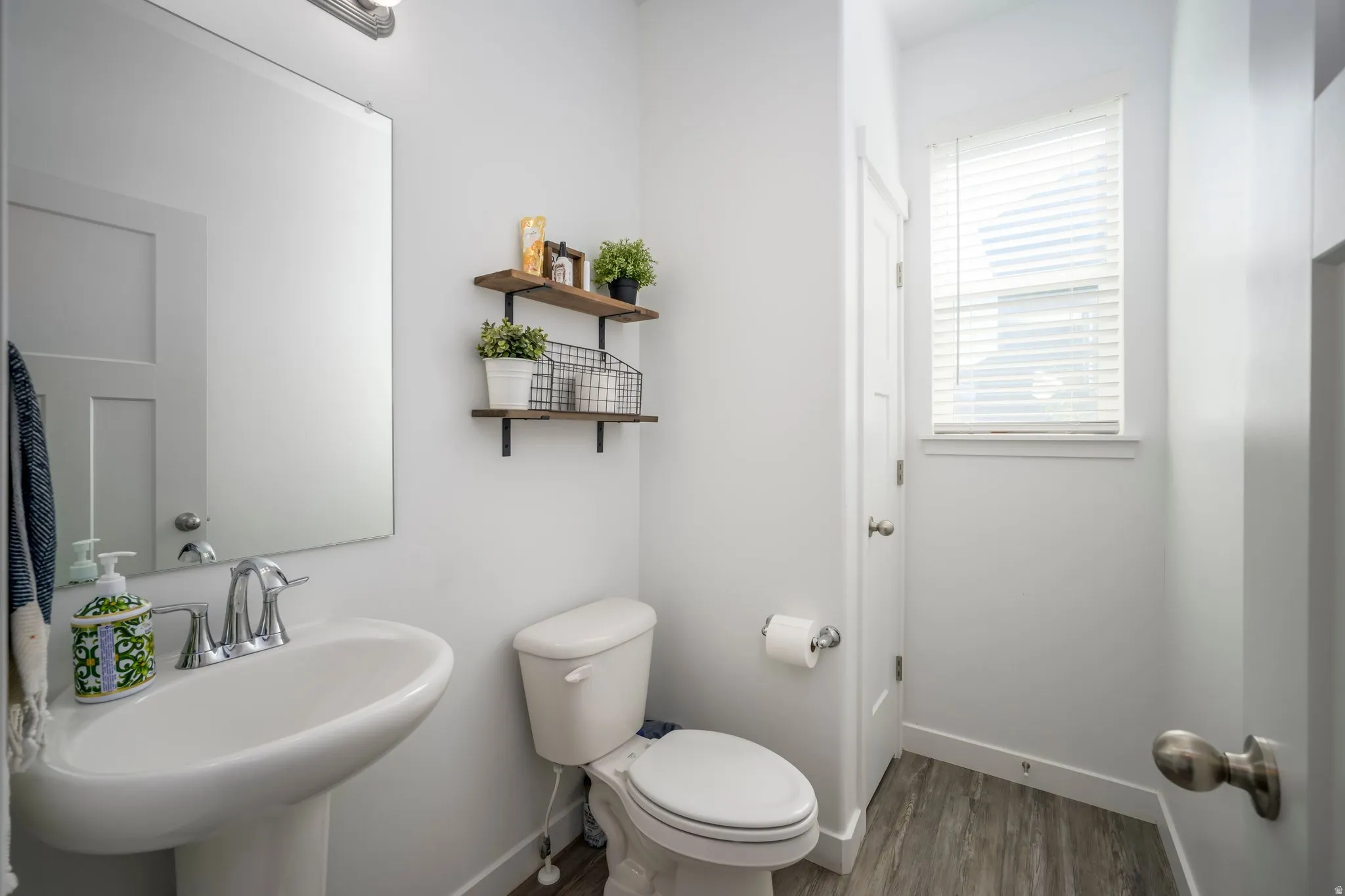 Compact bathroom featuring a pedestal sink with chrome faucet, a rectangular mirror, a window with blinds, and wood-finish flooring