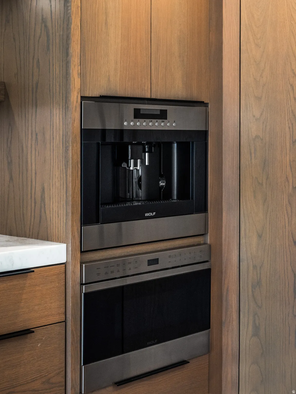 Integrated stainless steel built-in coffee system with a complementing wall oven, set within custom wood cabinetry with black linear hardware and a white stone countertop