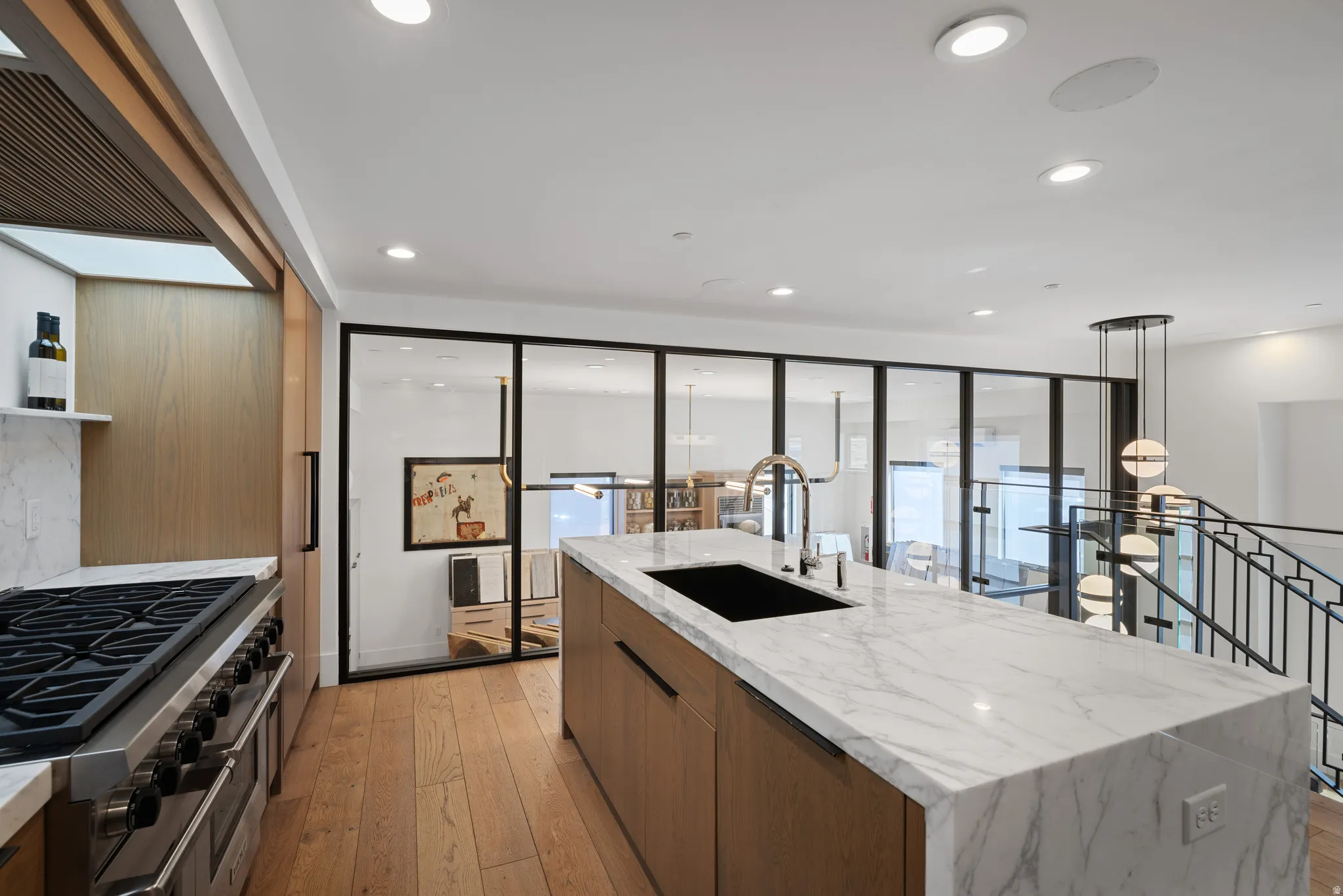 Spacious kitchen featuring a large waterfall island with integrated sink, stainless steel range with multiple burners, wood-finish flooring, and a contemporary pendant light fixture